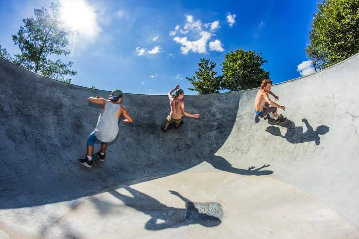 Trois enfants jouent dans un skatepark en faisant du skateboard sur une surface inclinée en béton, avec un ciel bleu et quelques nuages. Leurs ombres sont visibles sur la surface.