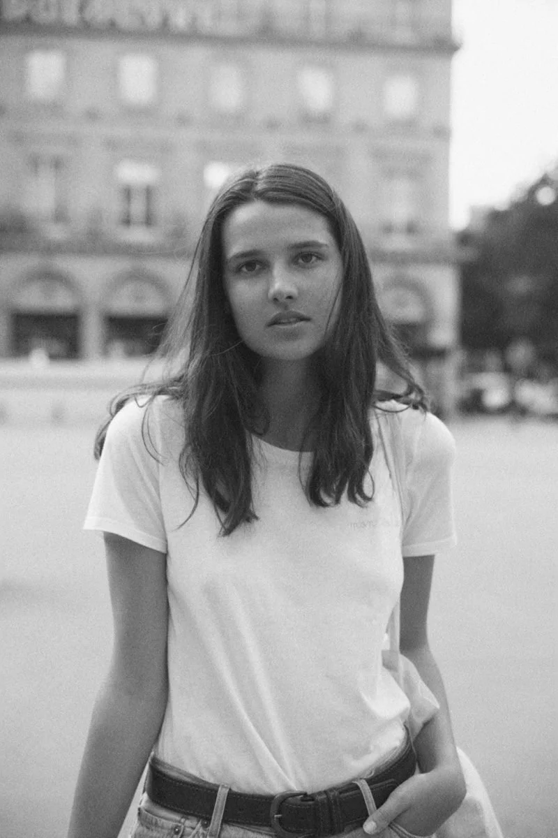 Une jeune femme à l'apparence décontractée, avec des cheveux longs et foncés, portant un t-shirt blanc et un jean, se tient devant un bâtiment en arrière-plan dans une scène en noir et blanc.