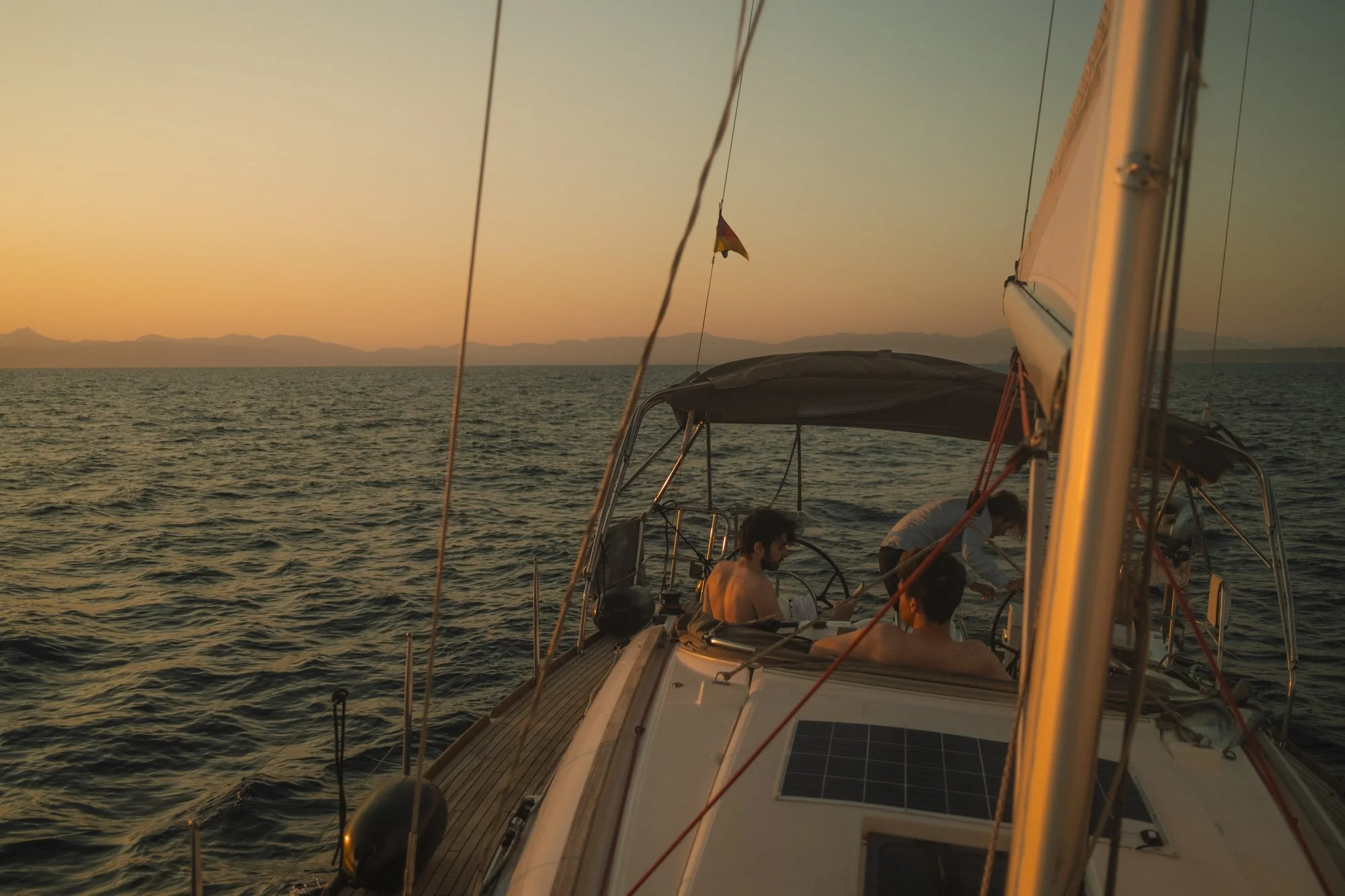 Une scène au coucher du soleil sur un bateau avec plusieurs personnes à bord, vue sur la mer et une chaîne de montagnes à l'horizon, avec un drapeau flottant dans le vent à majorque