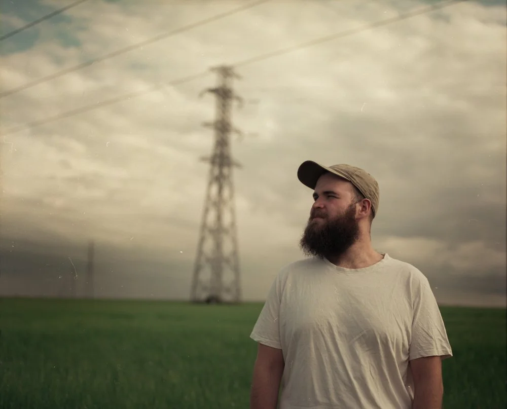 Homme avec une barbe portant un T-shirt beige et une casquette dans un champ vert, avec une tour électrique en arrière-plan, sous un ciel nuageux.