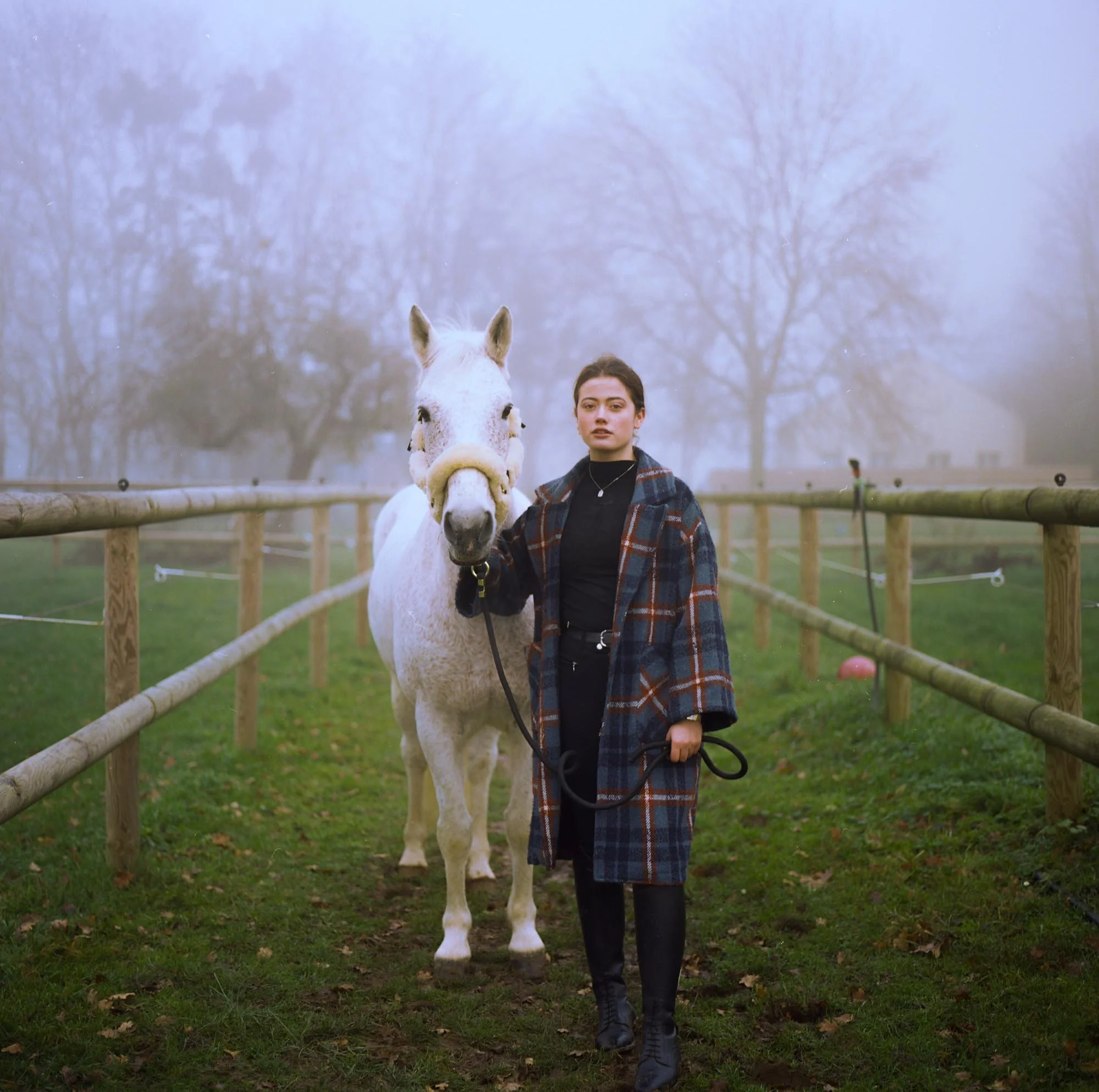Une jeune femme debout à côté d'un cheval blanc dans un enclos en bois, avec des arbres sans feuilles en arrière-plan par temps brumeux.