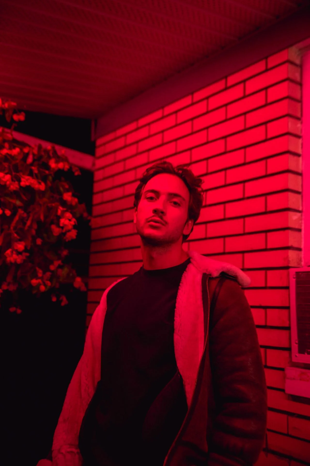 Un homme se tient devant un mur en briques, éclairé par une lumière rouge, avec des plantes à gauche.