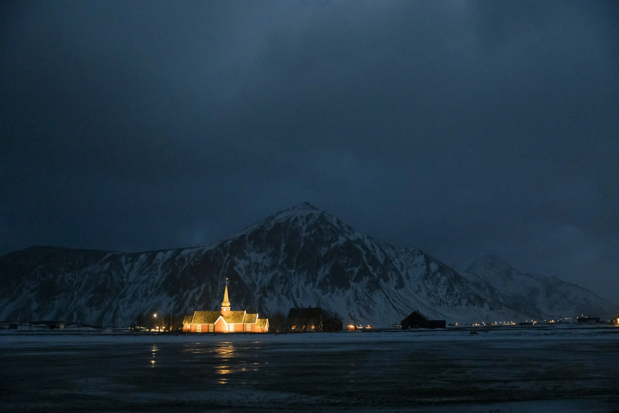Une église au centre d'un village avec une montagne enneigée en arrière-plan, de nuit, avec un ciel sombre et quelques lumières dans le village à Tromsø