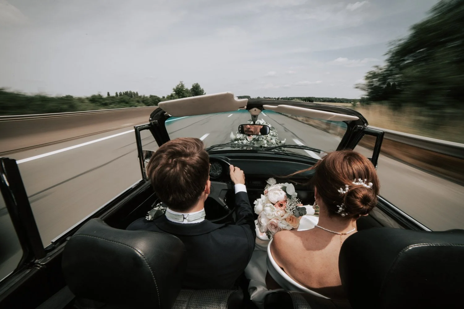Un couple marié conduit une voiture décapotable sur une route, la mariée tient un bouquet de fleurs et porte une robe de mariage, pendant qu'ils se dirigent vers leur lune de miel.