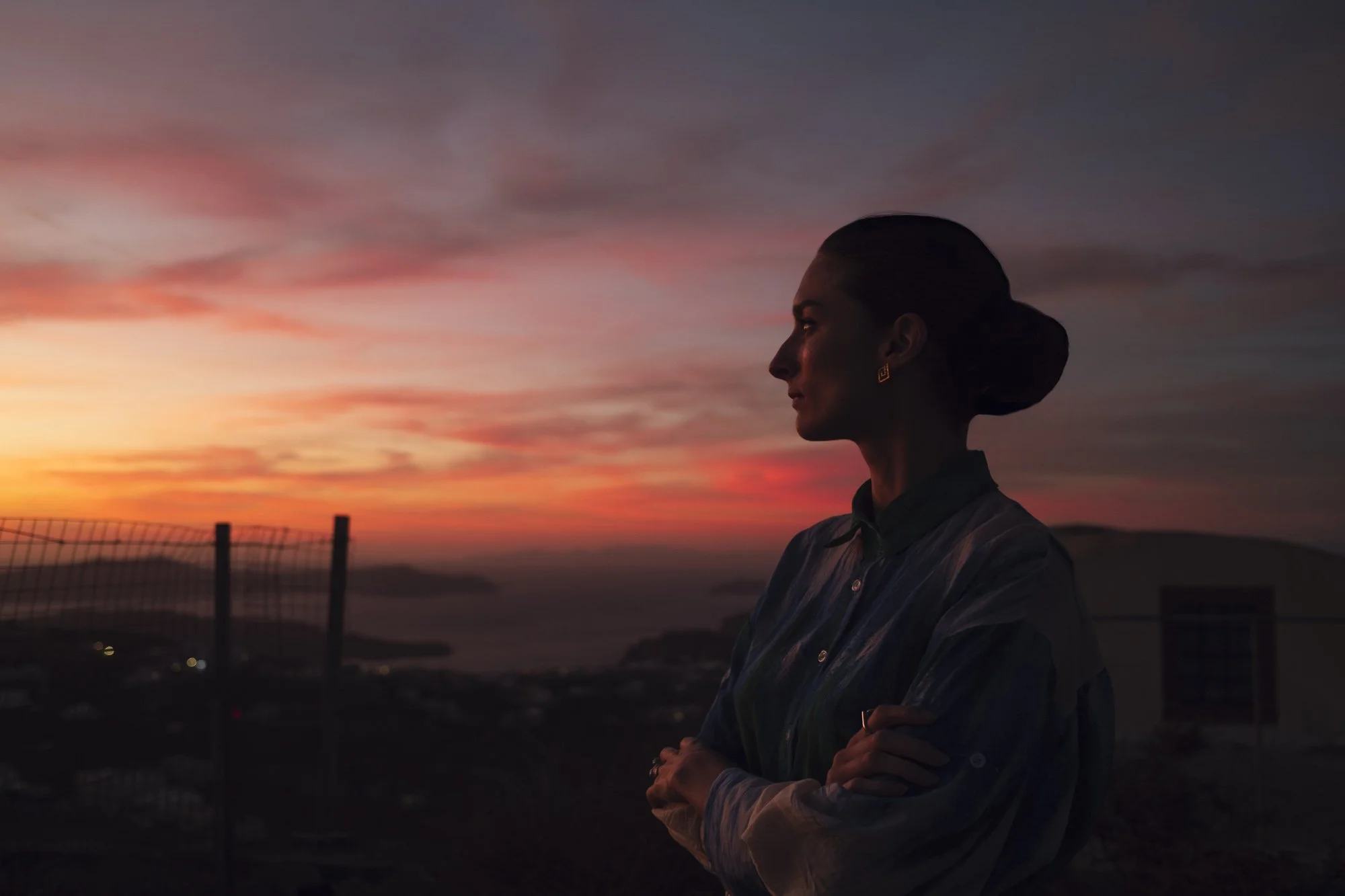 Une femme en profil, les bras croisés, regarde le coucher de soleil avec un ciel aux couleurs roses et violettes, en arrière-plan un paysage urbain et des structures de pont à santorin