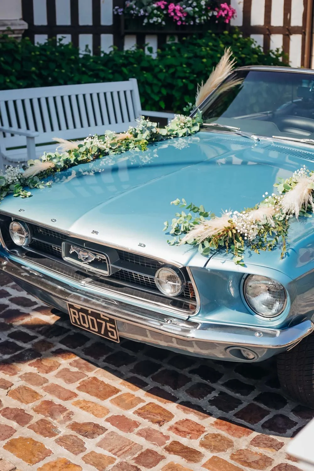 Voiture ancienne de couleur bleu clair décorée de fleurs pour un mariage, stationnée sur un pavé en briques, avec une clôture en bois et un banc blanc en arrière-plan.
