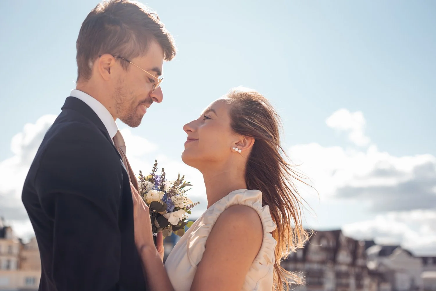 Un couple s'apprête à s'embrasser lors d'une cérémonie en plein air, la femme porte une robe beige et des boucles d'oreilles, l'homme porte un costume noir, l'homme tient un bouquet de fleurs.