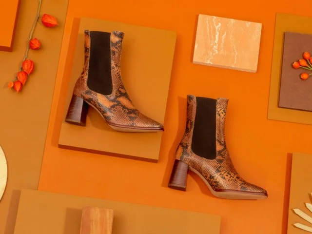 Une paire de bottines à talons en tissu imitation serpent sur un fond orange, entourée de divers objets décoratifs. Sarenza