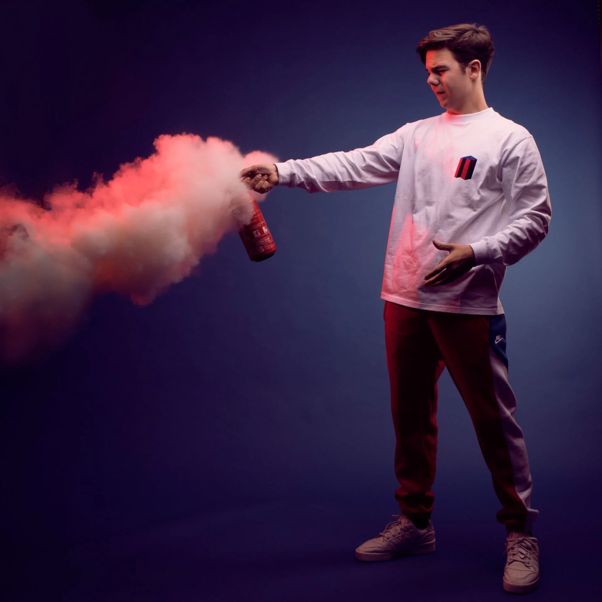 Un jeune homme blanc avec des vêtements de sport déployant un extincteur, libérant de la fumée colorée rose et blanche, dans un fond foncé.