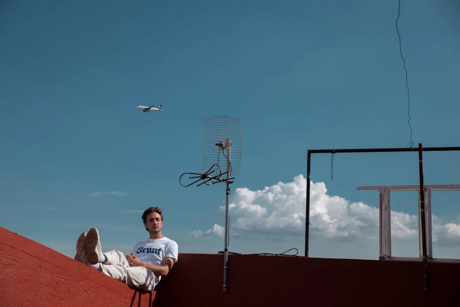 Jeune homme assis sur un toit rouge avec des appareils d'antenne et un avion dans le ciel derrière lui.