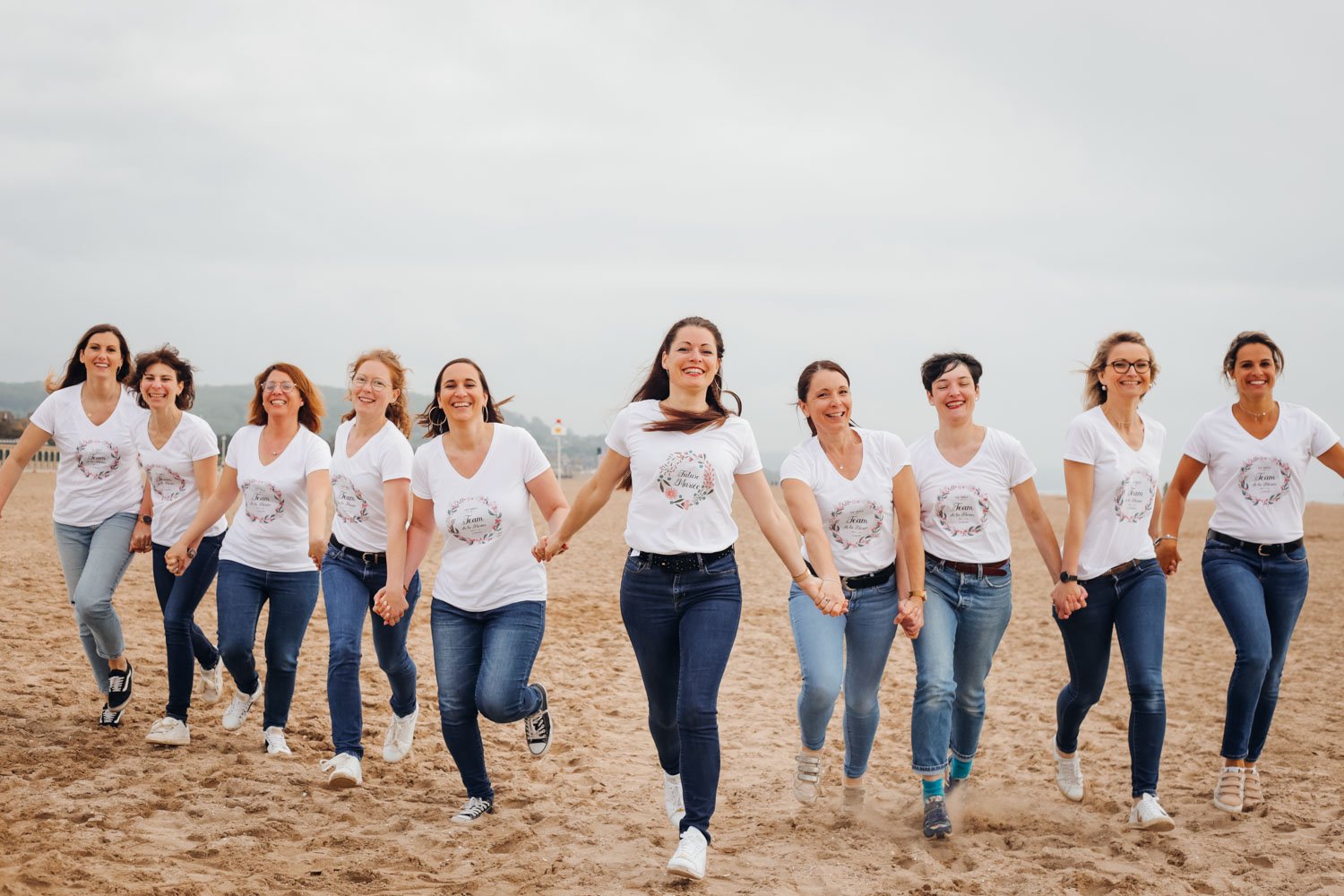Groupe de neuf femmes tenant des mains en marchant sur la plage, toutes portant des t-shirts blancs avec un design floral, souriantes, avec un ciel nuageux en arrière-plan.
