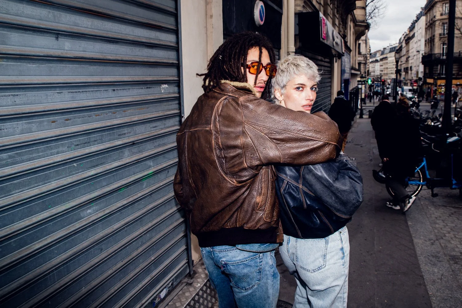 Deux jeunes femmes en tenues décontractées près d'une boutique fermée dans une rue urbaine, l'une porte des lunettes de soleil orange, l'autre un manteau noir.