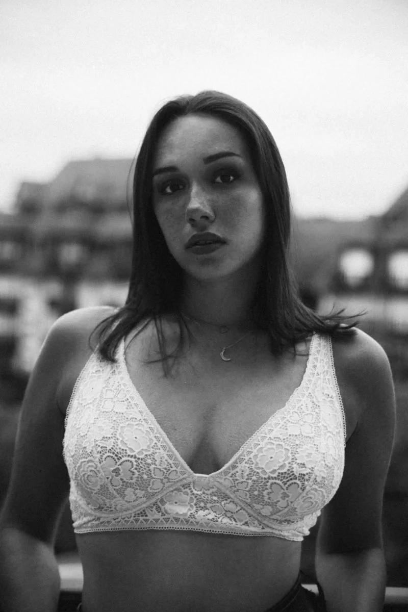 Une jeune femme avec des cheveux bruns et une expression sérieuse, portant un haut en dentelle blanche et un petit collier pendentif en forme de lune, debout devant un fond flou de bâtiments