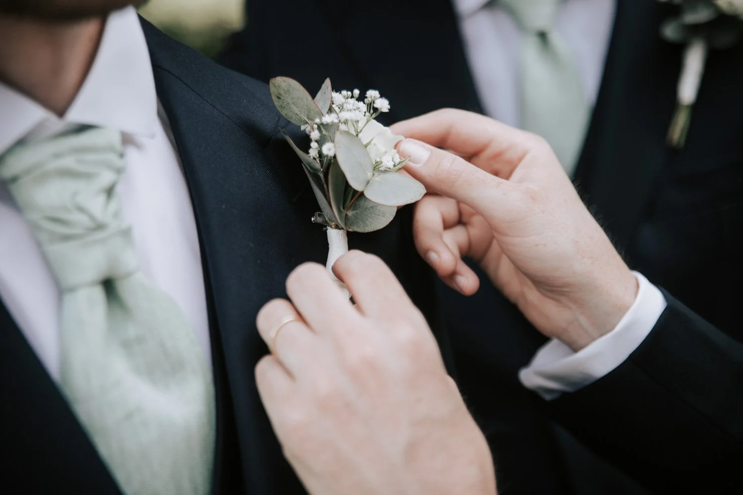 Une personne aide à fixer un boutonnière de fleurs blanches et de feuillage sur le costume noir d'une autre personne lors d'une cérémonie de mariage.
