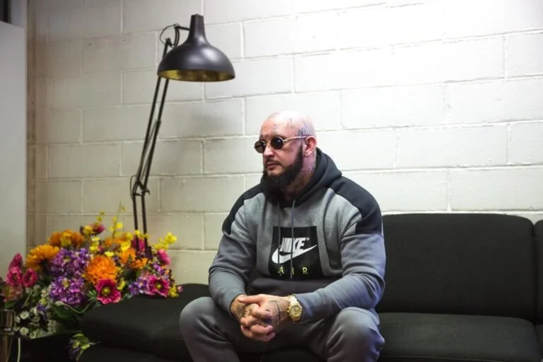 Un homme assis sur un canapé noir dans une pièce avec un mur blanc, portant des lunettes de soleil, un hoodie gris avec un logo Nike, une montre en or et une barbe, à côté d'un lampadaire et d'un bouquet de fleurs colorées.