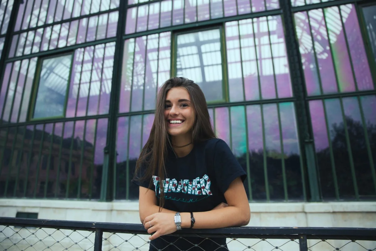 Une jeune femme souriante pose devant un bâtiment en verre avec des reflets violets.
