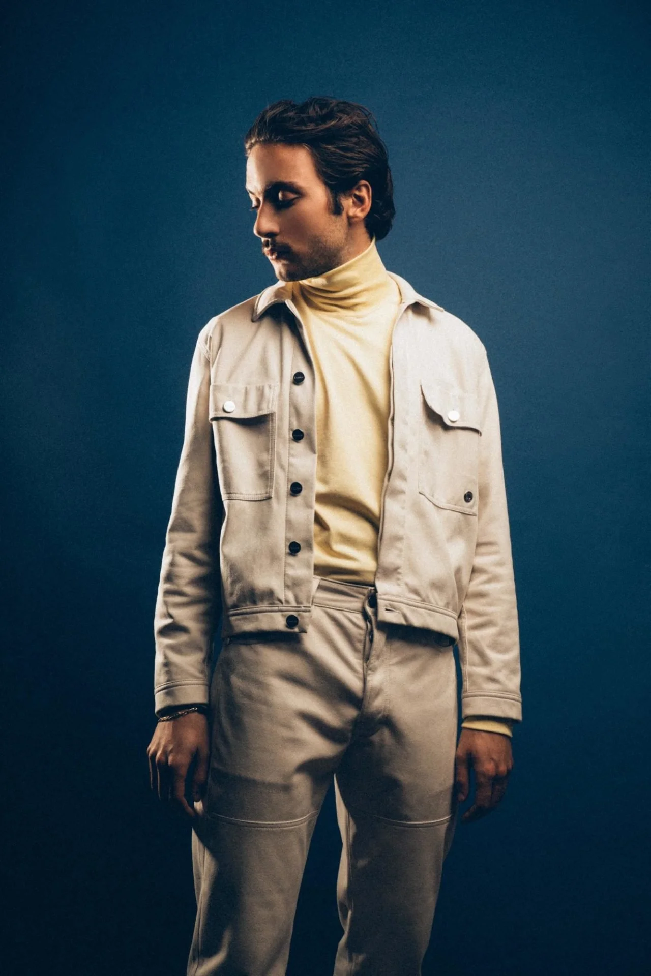 Jeune homme en vêtements beige, debout contre un fond bleu foncé, regardant vers le bas.