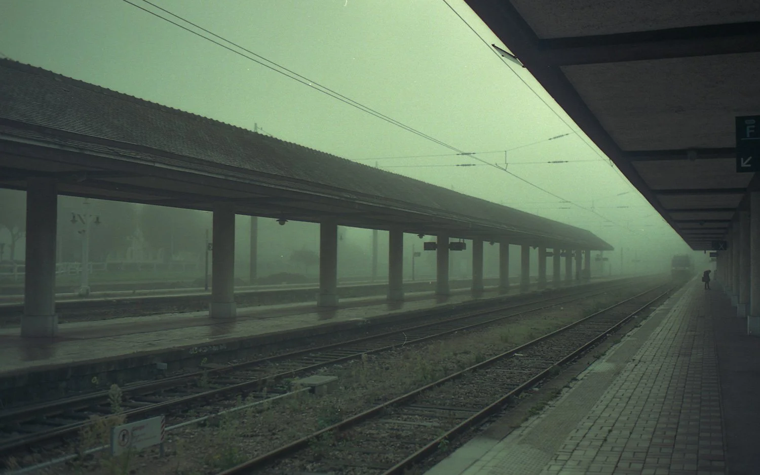 Une gare ferroviaire sous un brouillard épais, avec des trains à l'horizon et une personne avec un parapluie sur le quai