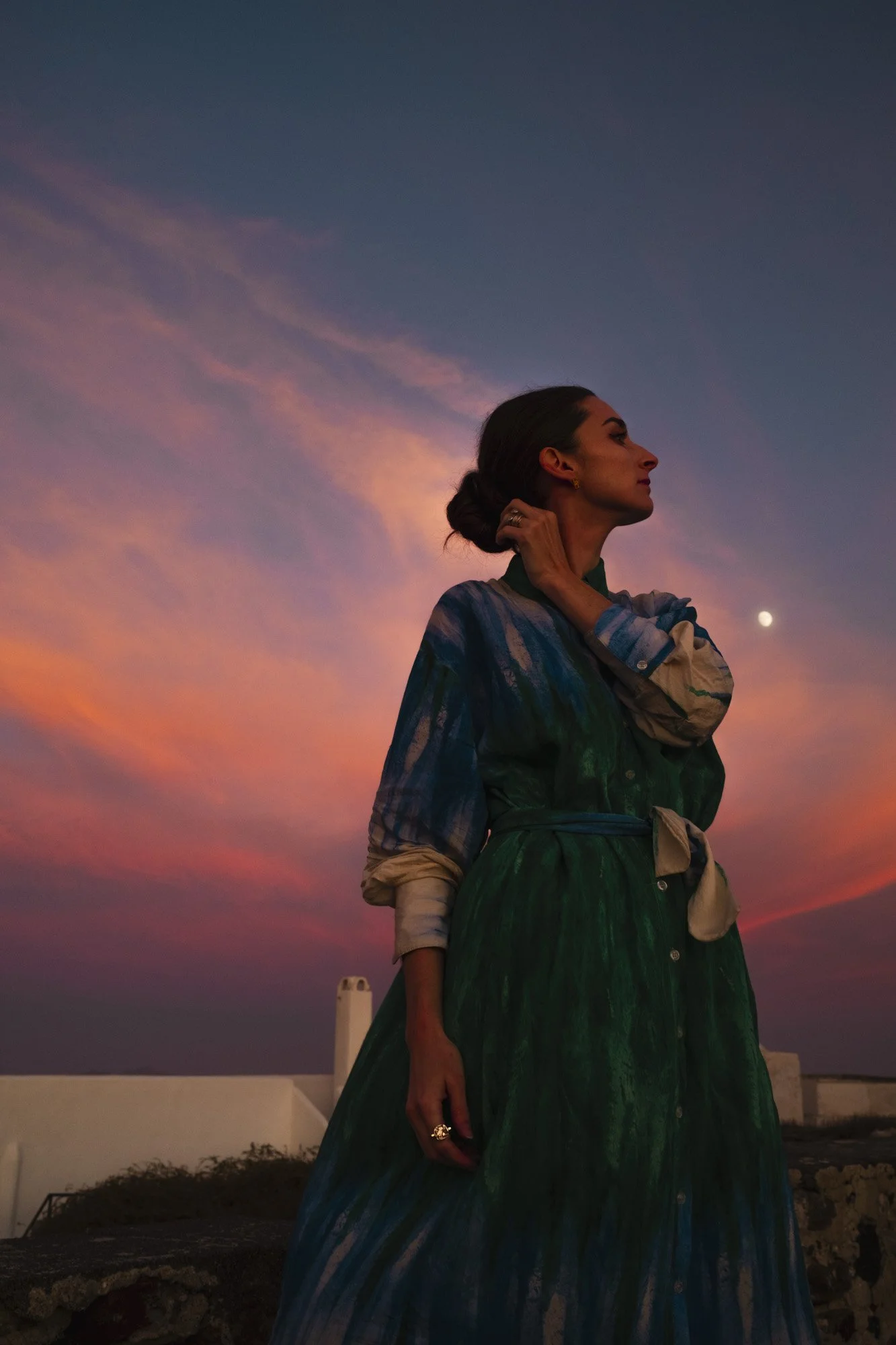 Une femme debout sur un cadre en pierre, portant une robe verte avec des manches longues, sous un ciel coloré au coucher du soleil, avec la lune en arrière-plan à santorin