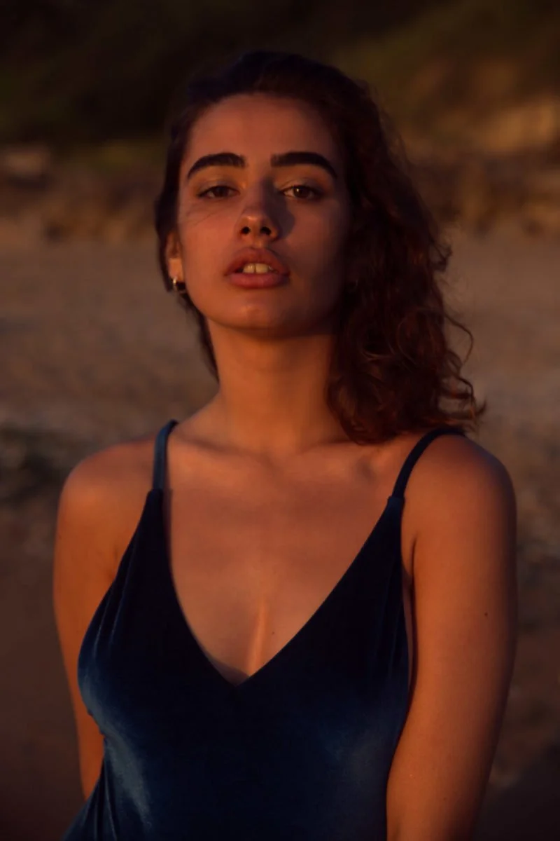 Une jeune femme aux cheveux bouclés et aux sourcils épais, portant un haut noir, se tient sur une plage au coucher du soleil, regardant directement l'appareil photo.