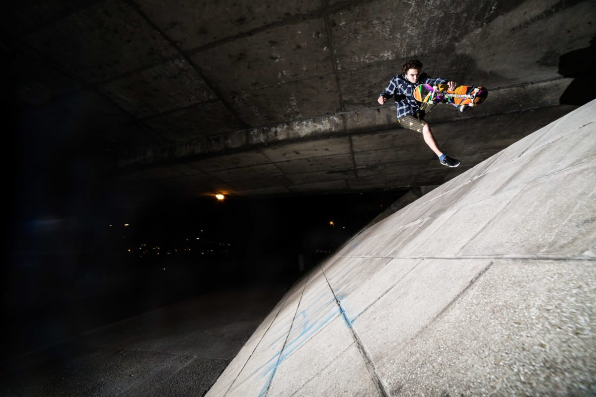 Un jeune homme en skateboard effectue un trick sur un mur en béton sous un pont la nuit.