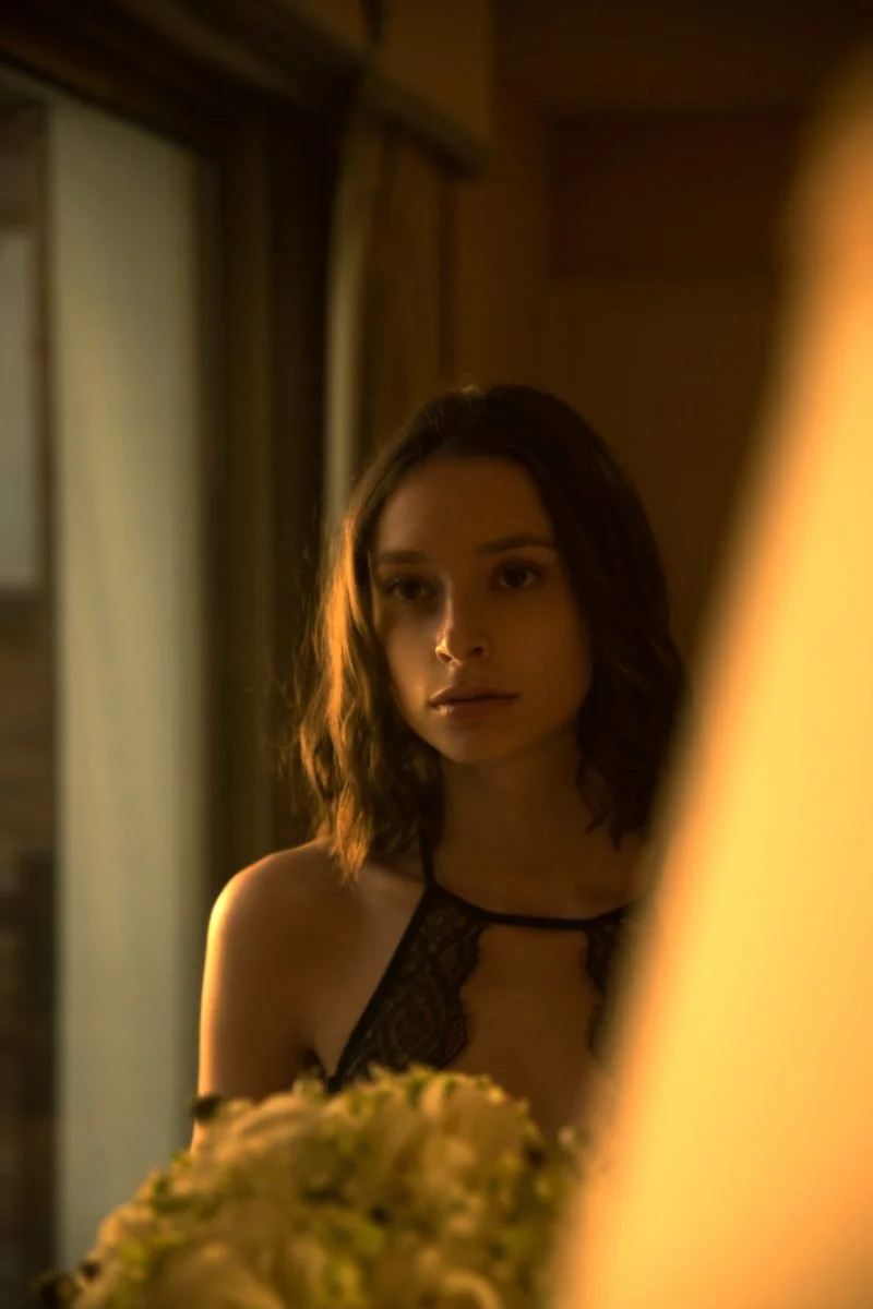 Une jeune femme en lingerie noire regarde dans un miroir dans une pièce avec une ambiance chaleureuse.