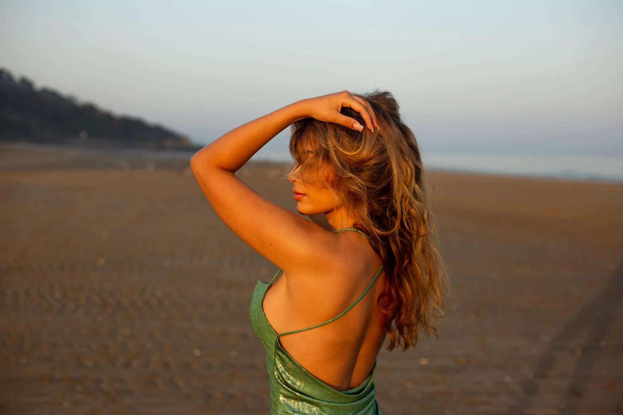 Une femme aux cheveux roux ondulés se tient sur une plage au coucher du soleil, portant une robe verte en satin, avec la mer et une falaise en arrière-plan.