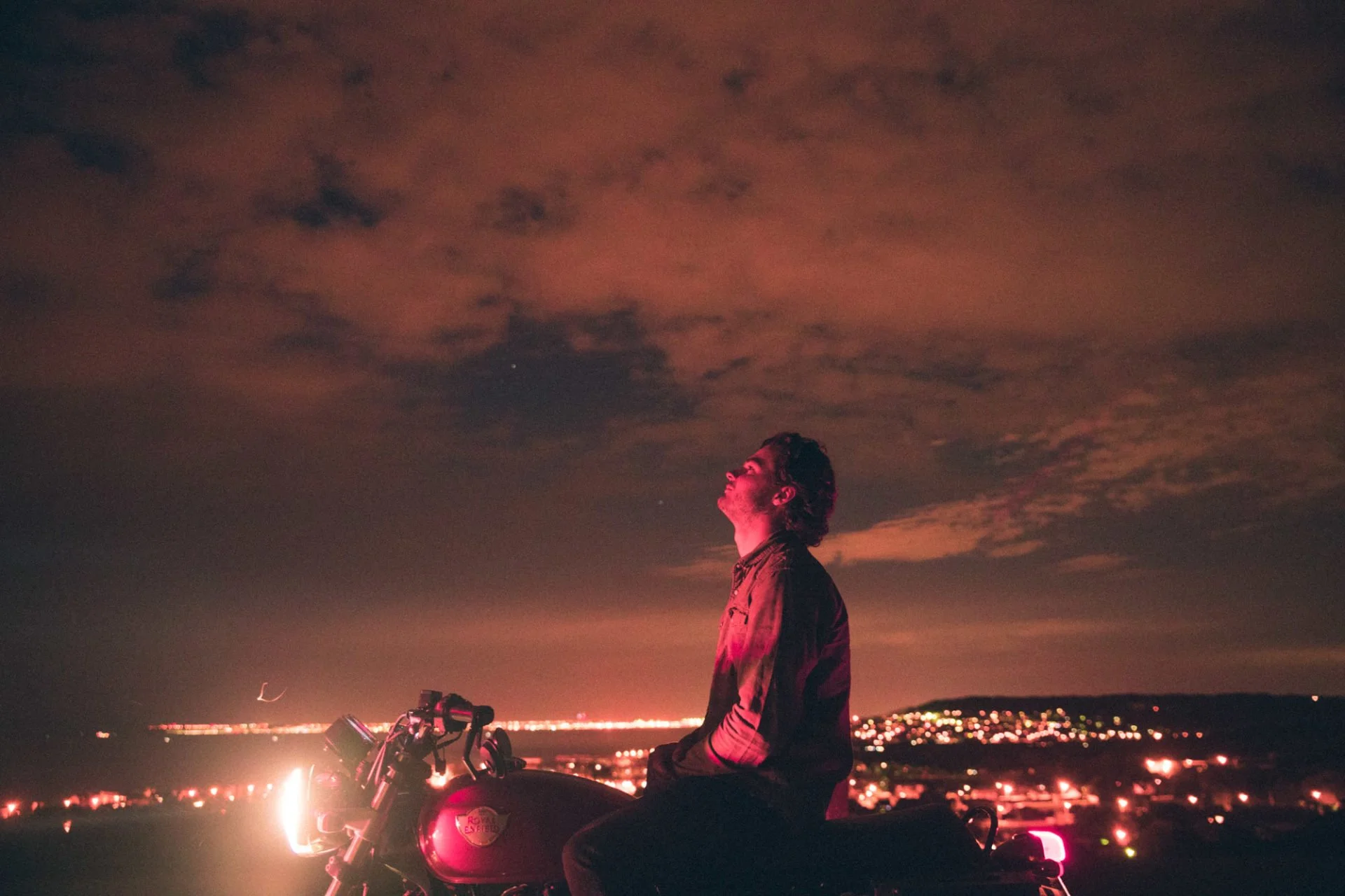 Un homme en veste regardant le ciel nocturne avec des nuages, proche d'une moto sur une colline avec vue sur une ville illuminée en contrebas.