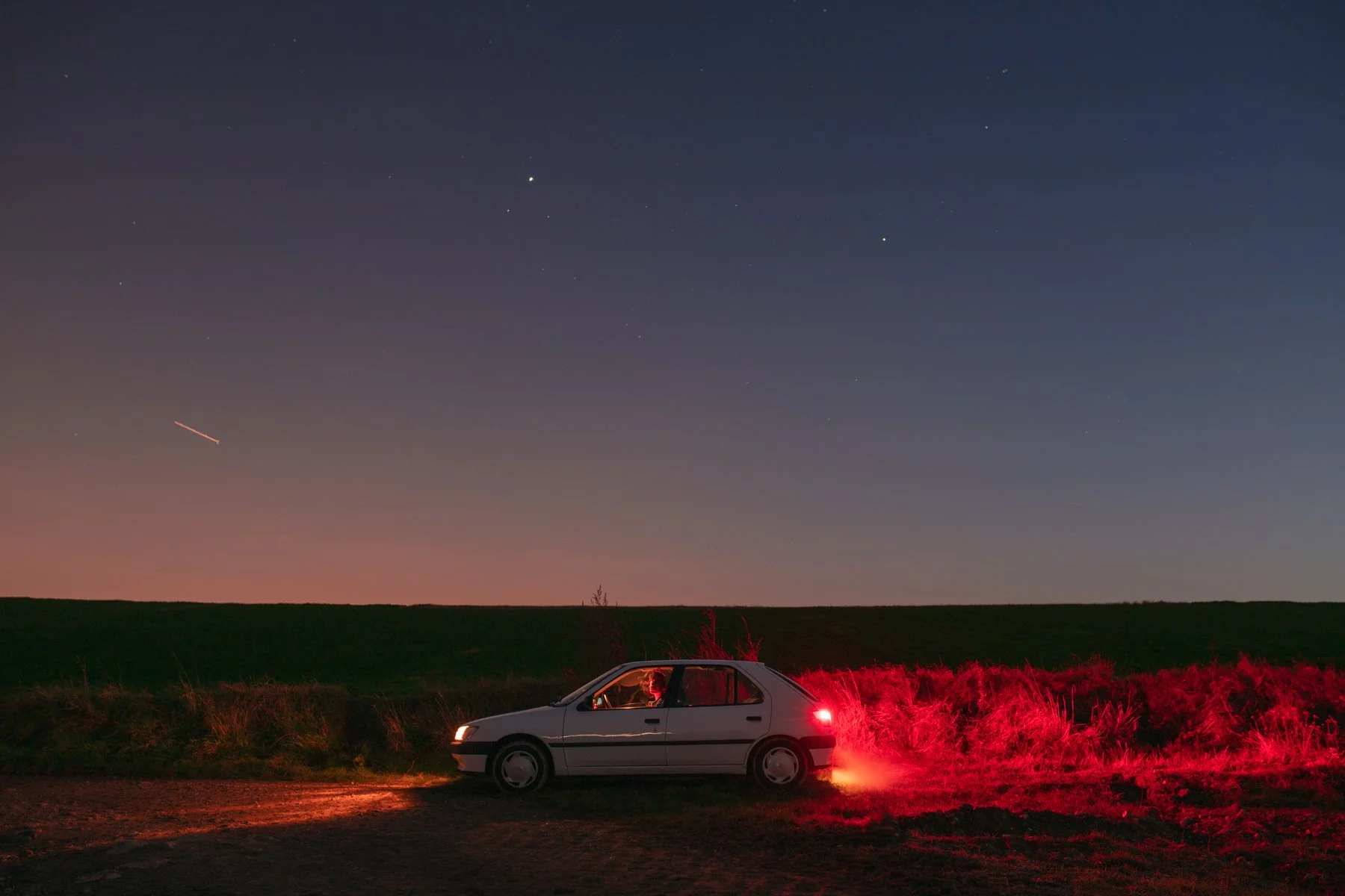 Une voiture blanche stationnée sur le bord d'une route dans un paysage rural, sous un ciel étoilé avec des étoiles brillantes et une traînée lumineuse d'un objet en mouvement.