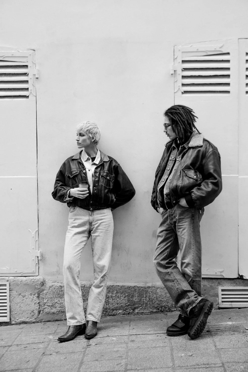 Deux personnes posent contre un mur blanc, portant des vestes en cuir, l'une avec des cheveux blonds courts et l'autre avec des dreadlocks, dans une scène en noir et blanc.