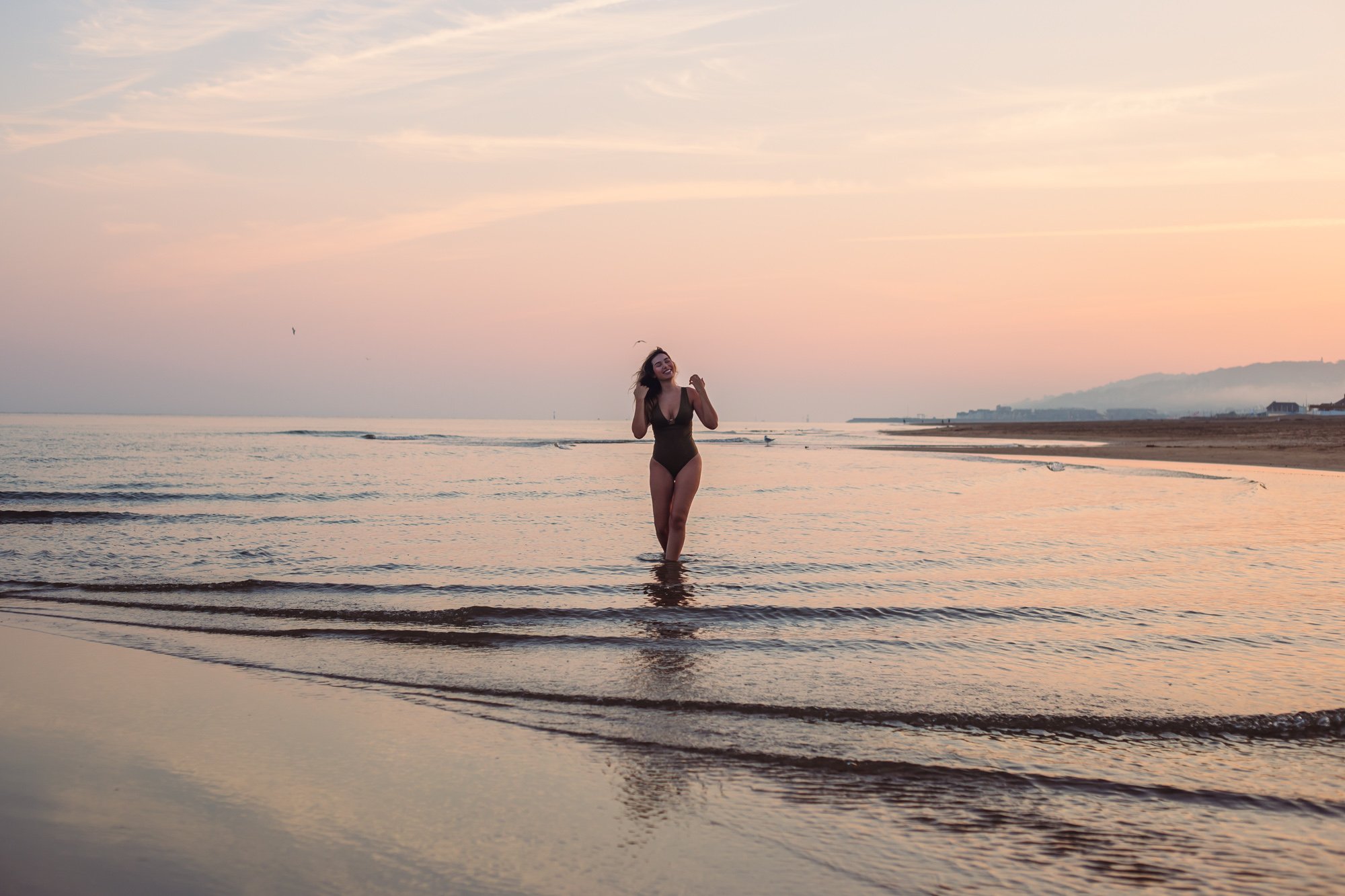 Femme souriante en maillot de bain dans la mer au coucher du soleil, paysages rocheux en arrière-plan.