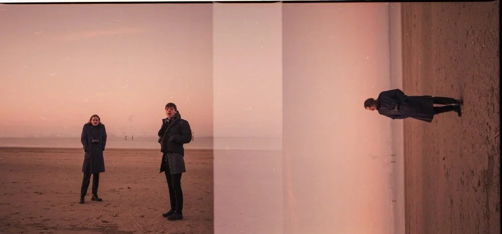Trois personnes en manteaux longs sur une plage déserte au coucher du soleil, avec un horizon lointain et un ciel rose pastel.