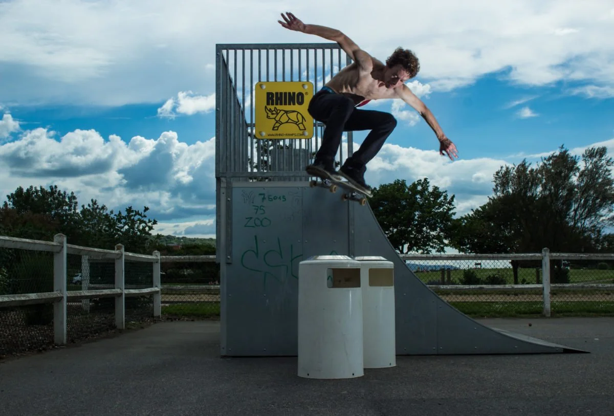 Un jeune homme sans t-shirt faisant du skate sur un repère à skate dans un parc, avec un ciel nuageux en arrière-plan.