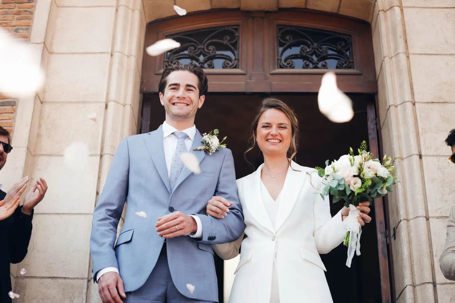 Un couple souriant sortant de l'église lors d'un mariage, la femme tenant un bouquet de fleurs pâles.