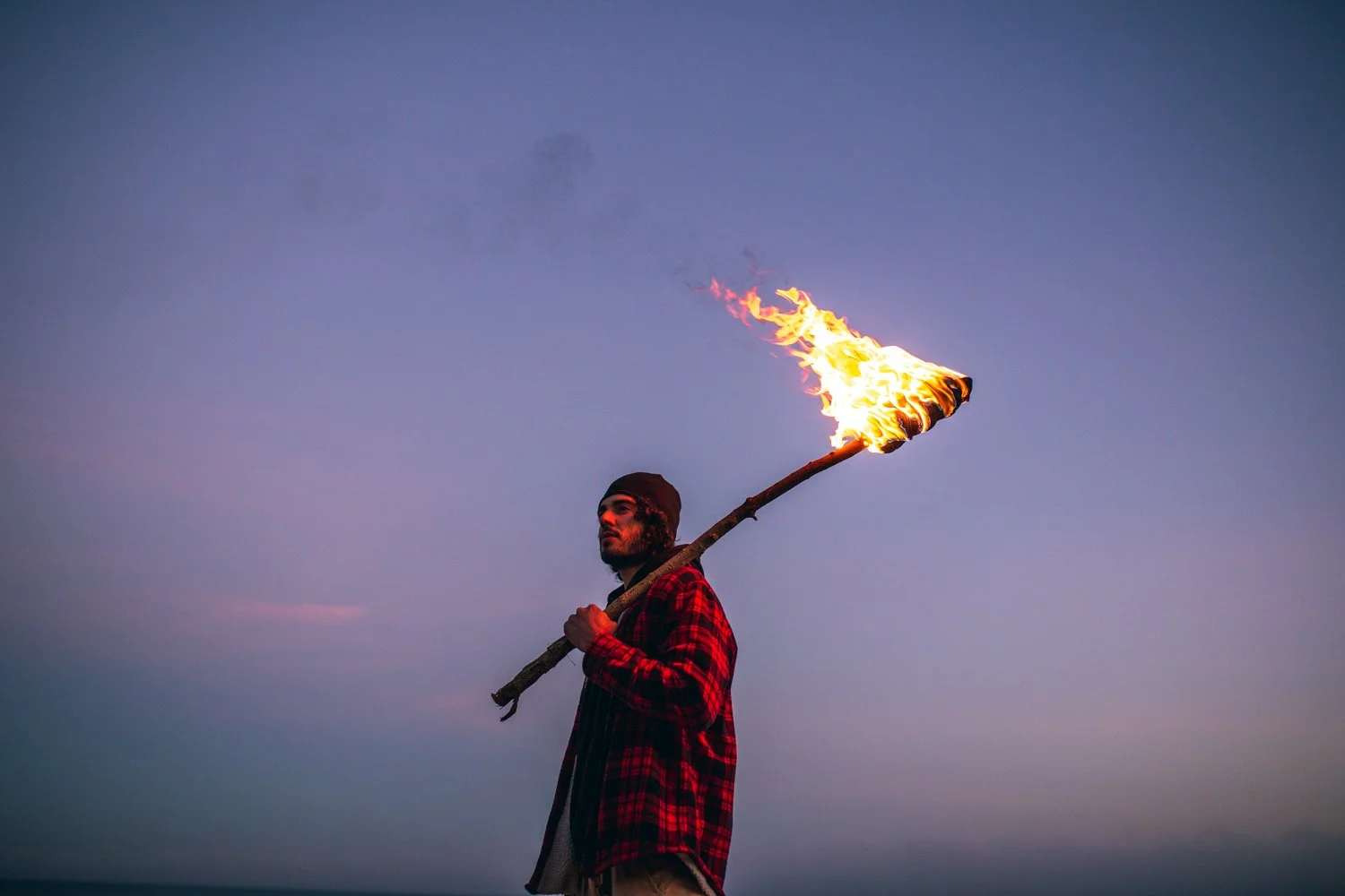 Un homme debout sur la plage tenant un bâton avec une flamme allumée à l'extrémité, ciel en crépuscule ou au lever du soleil.