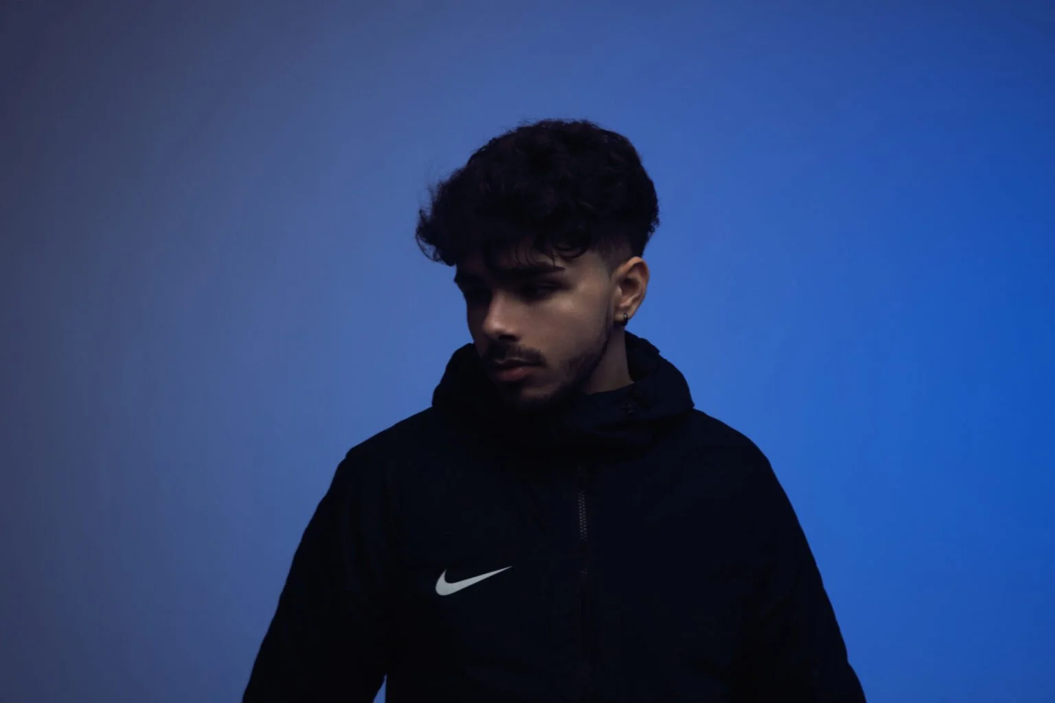 Jeune homme avec des cheveux foncés et une barbe, portant un sweat noir avec un logo Nike, regardant vers le bas, sur un fond bleu.