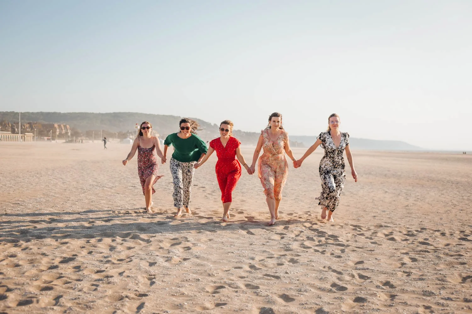 Cinq femmes marchant main dans la main sur une plage ensoleillée, toutes en vêtements colorés et fleuris.