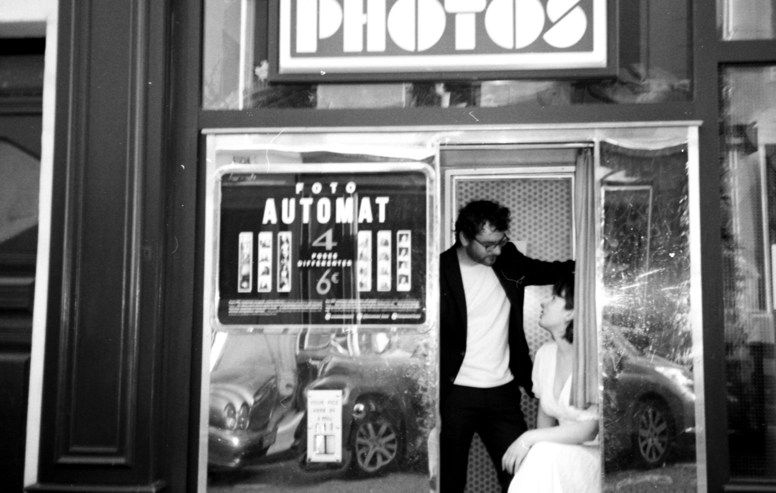 Deux personnes discutent devant un appareil photo automatique dans une cabine photo en noir et blanc.