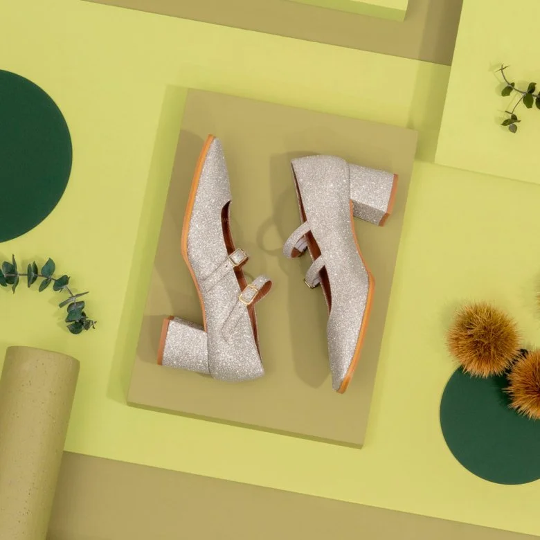Une paire de chaussures à talons hauts recouvertes de glitter argenté, affichée sur une surface beige, avec des éléments décoratifs verts et orange autour de l'image. Sarenza