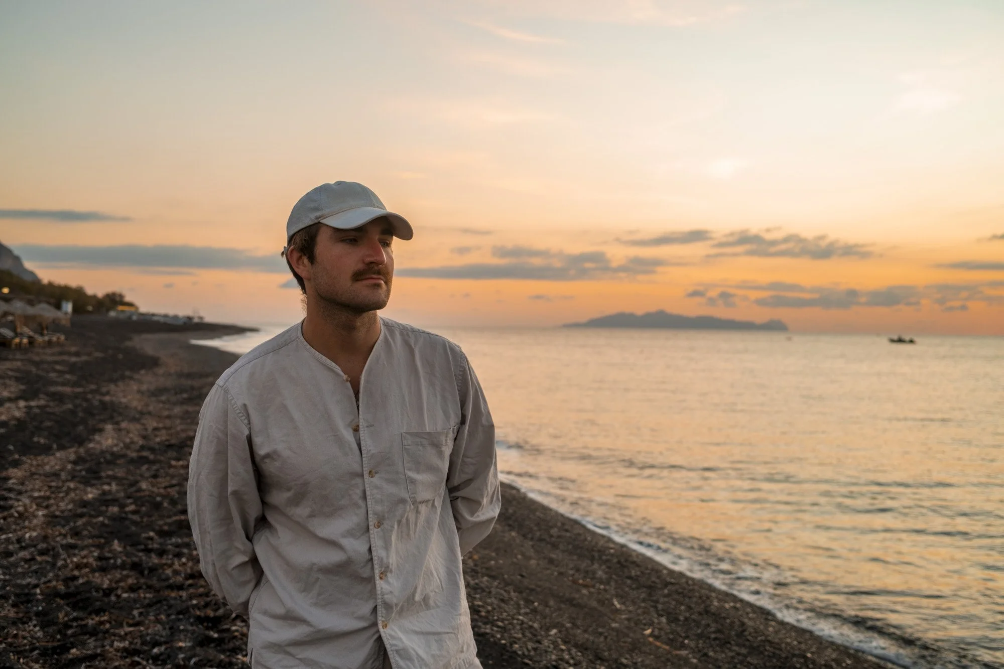 Un homme avec une casquette blanche et une chemise claire se tient sur la plage au coucher du soleil, regardant pensivement vers la mer. Encadré par un ciel orange et quelques nuages, avec des bateaux au loin et une montagne en arrière-plan.
