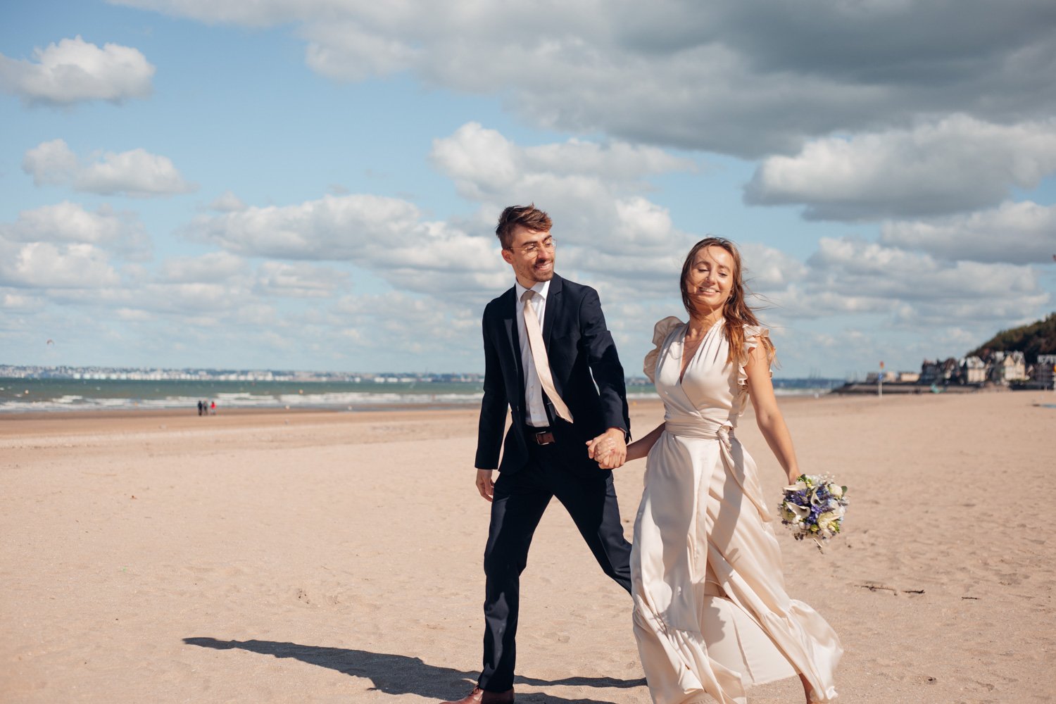 Un couple en vêtements de mariage marche sur la plage ensoleillée, tenant la main et souriant, avec le ciel nuageux en arrière-plan.