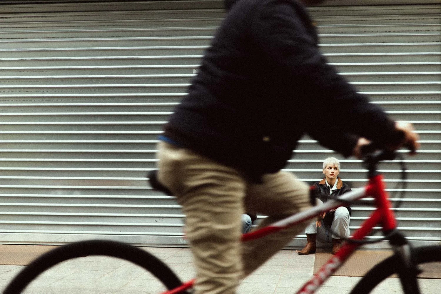 Une personne qui fait du vélo passe devant une femme assise contre un volet métallique fermé.