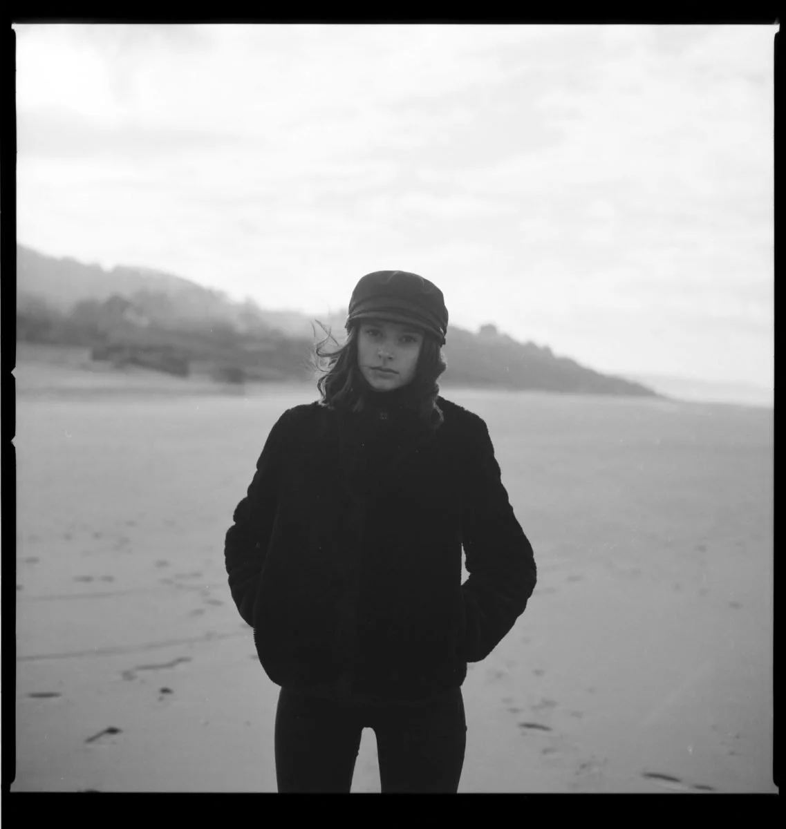 Une jeune fille en veste et chapeau regardant l'objectif sur une plage en noir et blanc.