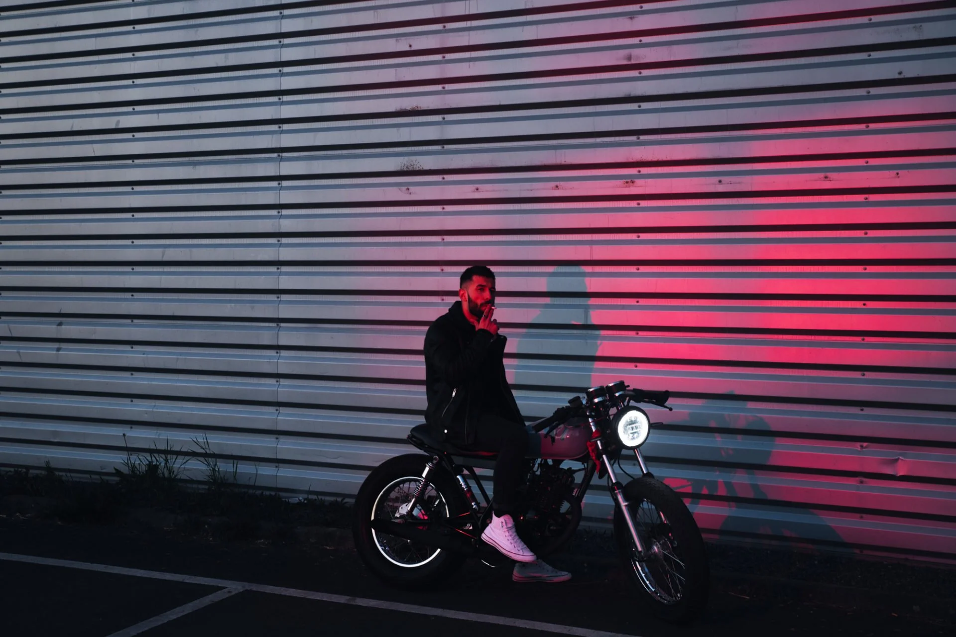 Un homme avec une barbe, portant une veste noire et des baskets blanches, assis sur un vélo devant un mur métallique avec des lignes horizontales, une lumière rouge éclaire le mur, créant une ombre de l'homme sur la surface.