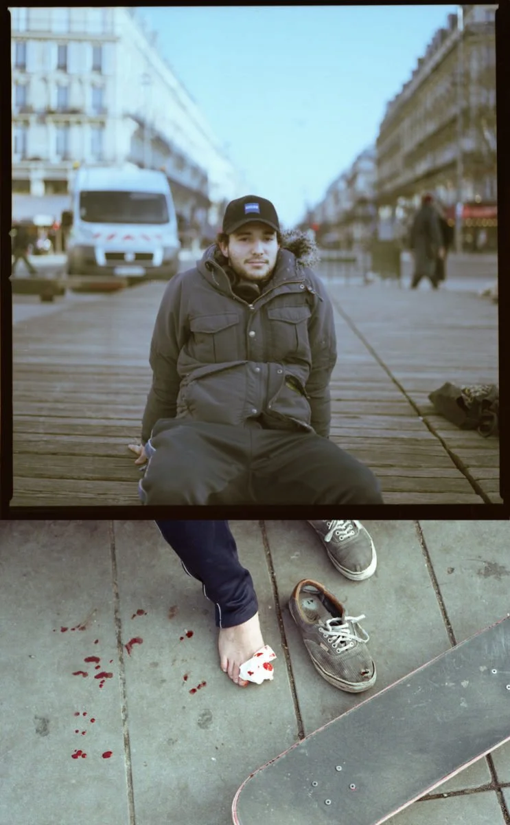 Un homme assis sur un quai en bois dans une rue urbaine, avec des bâtiments en arrière-plan, portant une veste noire et un bonnet, la tête légèrement inclinée vers l'avant. Devant lui, sur le sol en pierre, il y a une chaussure usée, une chaussure av