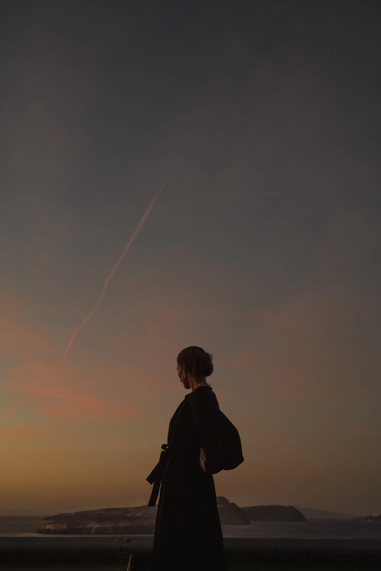 Une silhouette de personne habillée en noir regardant vers le coucher de soleil sur la mer, avec des falaises en arrière-plan et un ciel avec une traînée lumineuse à santorin