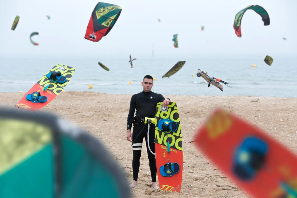 Un jeune homme debout sur la plage avec une planche de kite surf, entouré de plusieurs kite surfeurs dans le ciel.