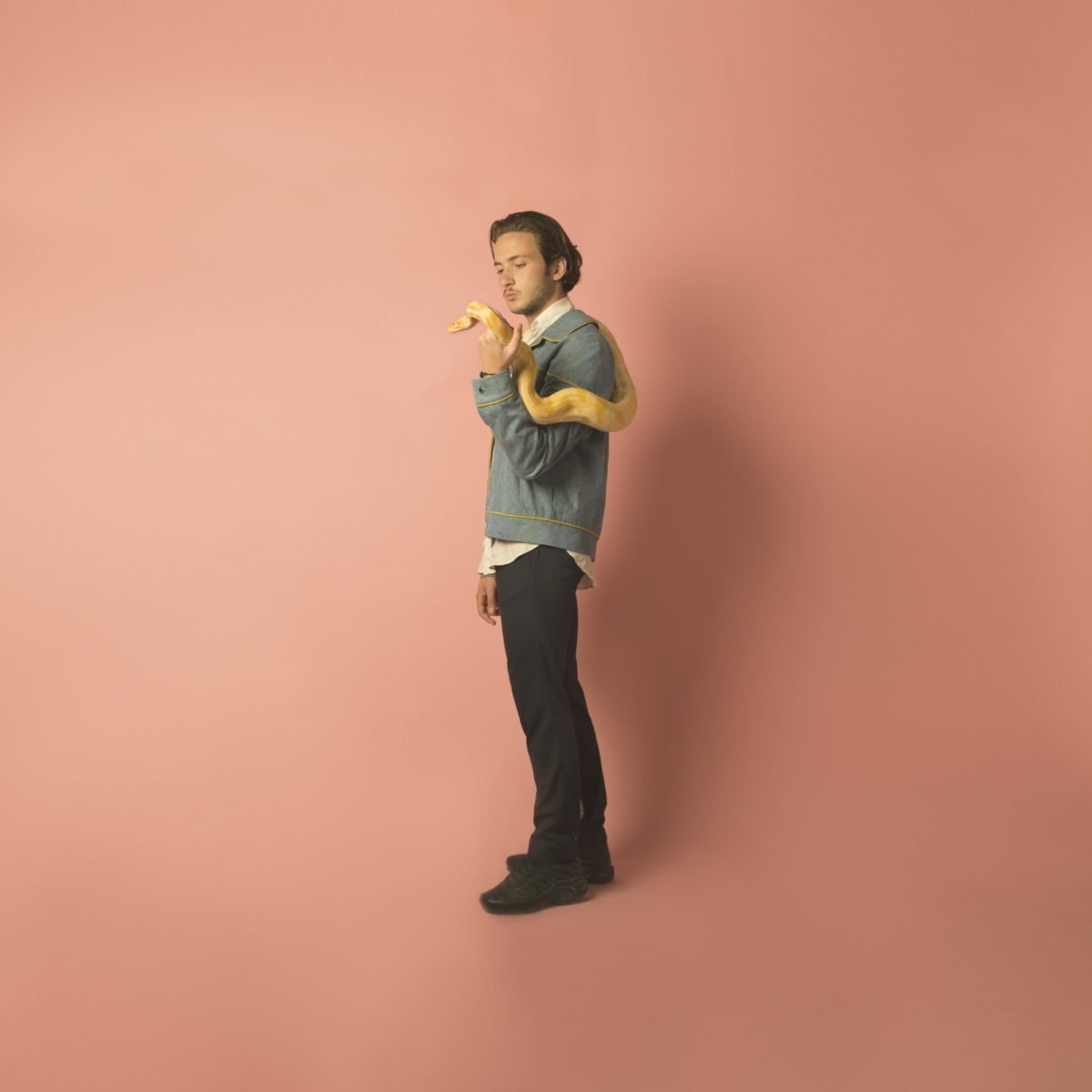 Jeune homme avec un serpent jaune enroulé autour de son bras, posture décontractée, fond rose pastel.