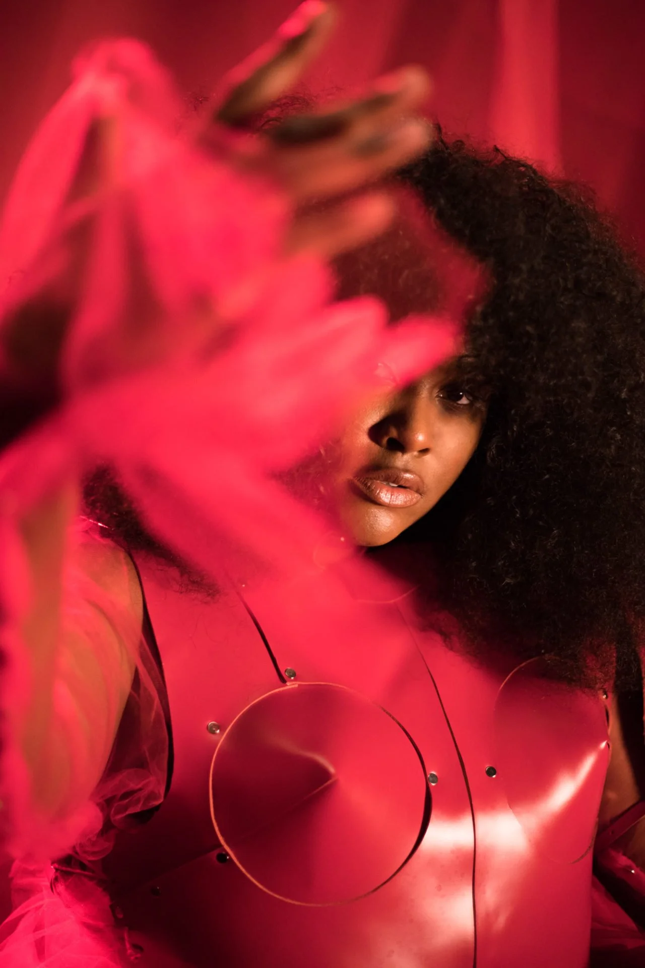 Une femme noire avec de cheveux volumineux et bouclés pose dans un éclairage rouge. Elle porte une matière brillante, probablement du latex, avec des cercles décoratifs.