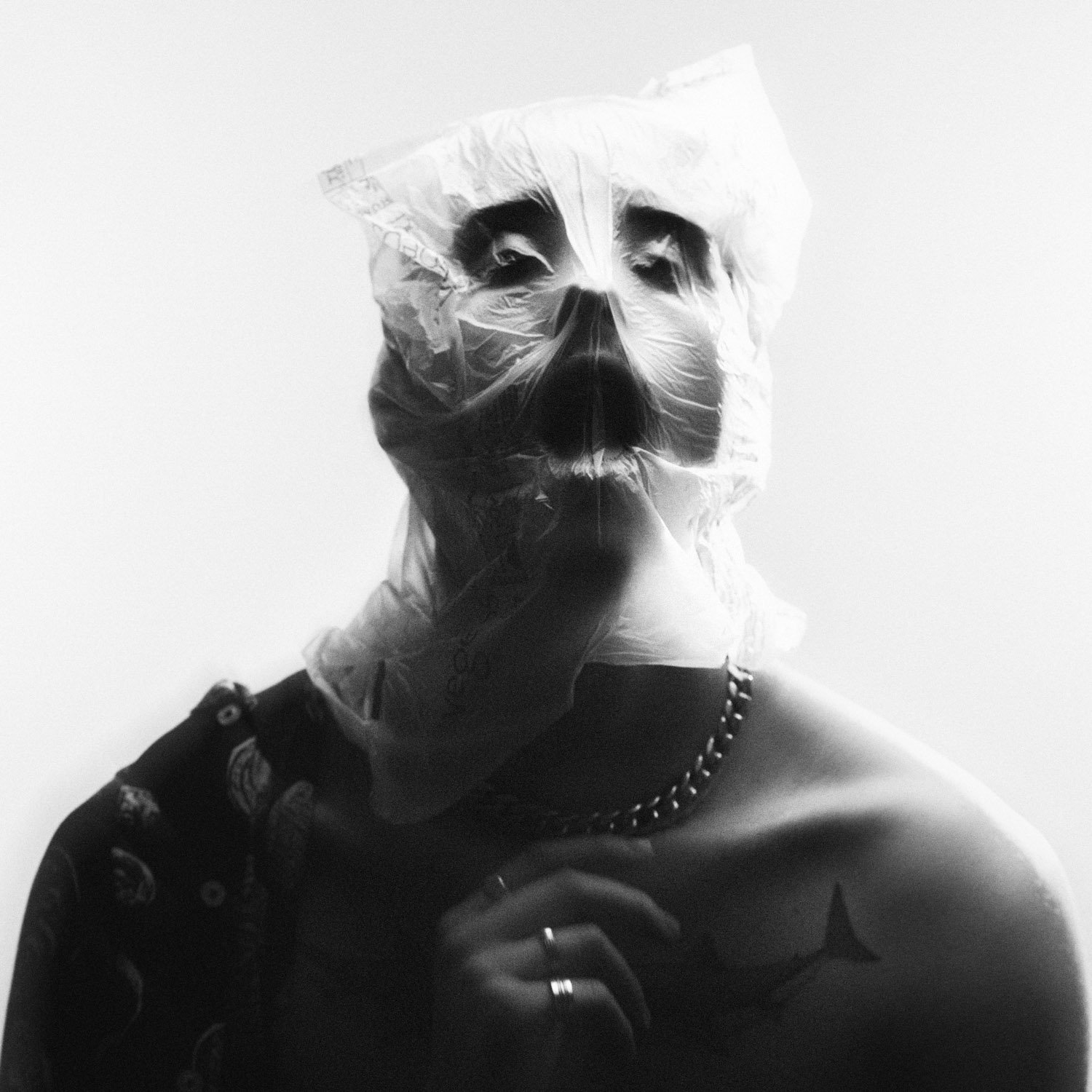 Image en noir et blanc d'une personne portant un masque en plastique, se rasant dans un miroir, avec un collier en chaîne, un tatouage d'étoile sur l'épaule, et des vêtements sombres.